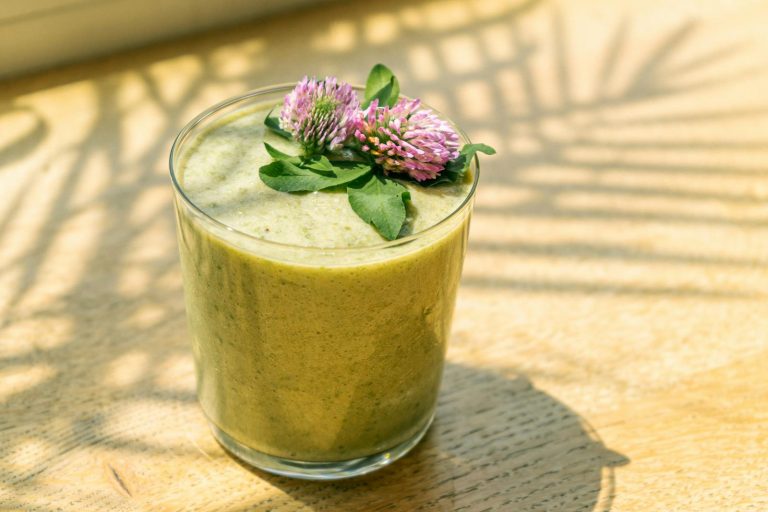 Close-up of a nutritious green smoothie topped with fresh purple clover flowers in sunlight.